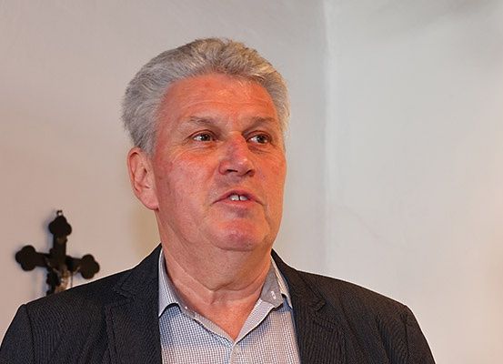 Ein älterer Mann mit grauem Haar und ernster Miene steht vor einer weißen Wand mit einem Kruzifix.