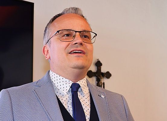 Ein Mann mit Brille und einem Westchen mit blau-weißem Karomuster lächelt vor einer Wand mit einem Kreuz.