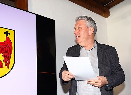 Ein Mann steht neben einem Bildschirm, der ein heraldisches Symbol zeigt, und hält ein Papier in der Hand, wahrscheinlich präsentiert er Informationen.