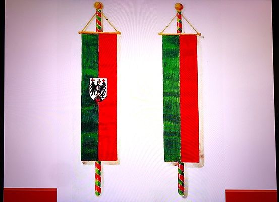 Zwei Flaggen mit grünem und rotem Hintergrund, die ein Wappen zeigen, hängen an einer weißen Wand.