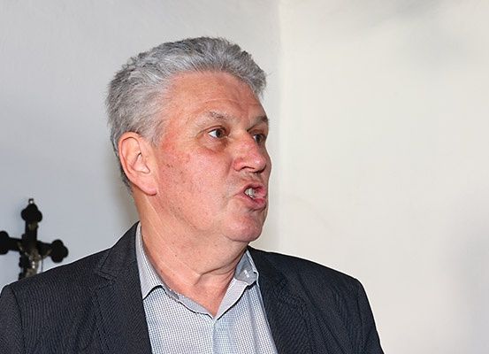 Ein älterer Mann mit grauem Haar, der einen schwarzen Anzug und ein graues Hemd trägt, steht vor einer weißen Wand mit einem Kruzifix.