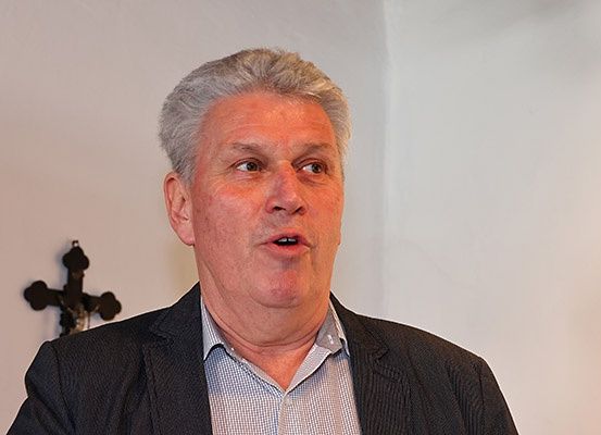 Ein Mann mit grauem Haar, der einen schwarzen Blazer und ein graues kariertes Hemd trägt, steht vor einer weißen Wand mit einem schwarzen Kreuz, das daran befestigt ist.
