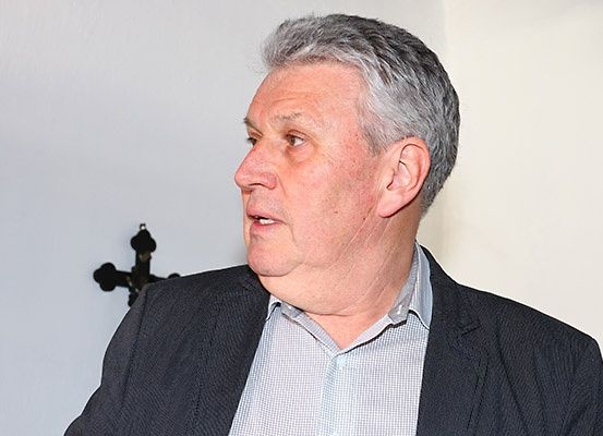 Ein älterer Mann mit grauem Haar steht und schaut nach links. Er trägt eine schwarze Anzugjacke über einem weißen karierten Hemd. An der Wand hinter ihm hängt ein schwarzes Kreuz.