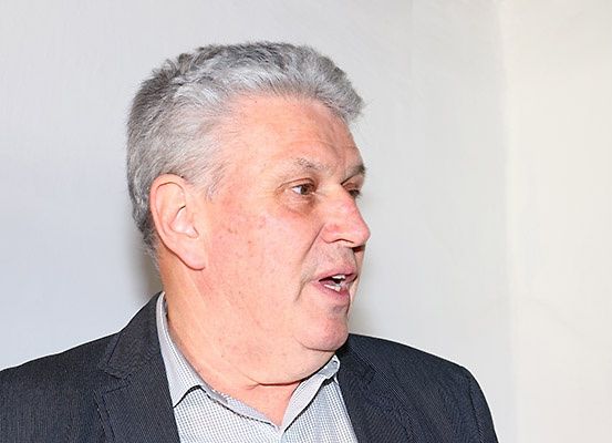 Ein älterer Mann mit grauem Haar steht und schaut nach rechts. Er trägt einen dunkelgrauen Blazer über einem weißen Hemd mit kleinem Kragen. Der Hintergrund ist eine schlichte weiße Wand.