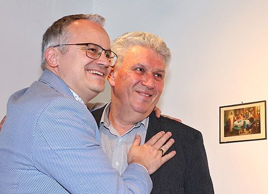 Zwei Männer in formeller Kleidung, einer mit Brille, lächeln und umarmen sich. Sie stehen neben einem gerahmten Bild an der Wand.