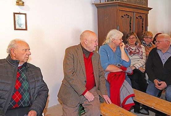 Eine Gruppe älterer Personen sitzt in einem Raum, vielleicht bei einer Gemeindeversammlung, und trägt Winterkleidung. Sie scheinen in ein Gespräch vertieft zu sein.