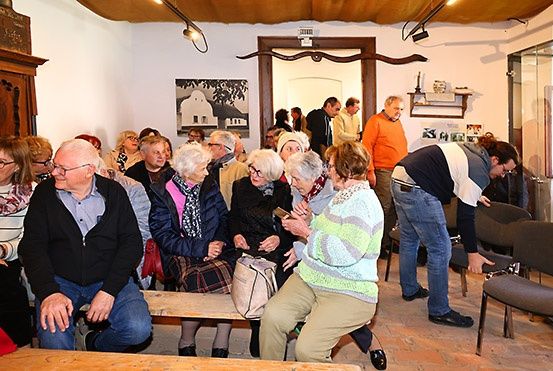 Eine Gruppe älterer Menschen sitzt zusammen in einem Raum, einige stehen und unterhalten sich. Es gibt Stühle, eine Holzbank und ein Bild an der Wand.