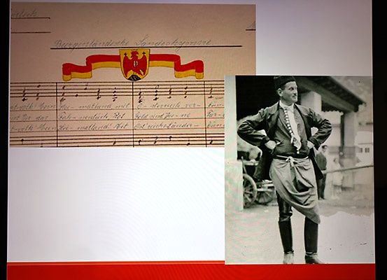 Ein Dokument mit Musiknoten und Text auf Deutsch, das ein Wappen zeigt. Ein Schwarz-Weiß-Foto eines Mannes in traditioneller Kleidung, der mit den Händen auf den Hüften steht.