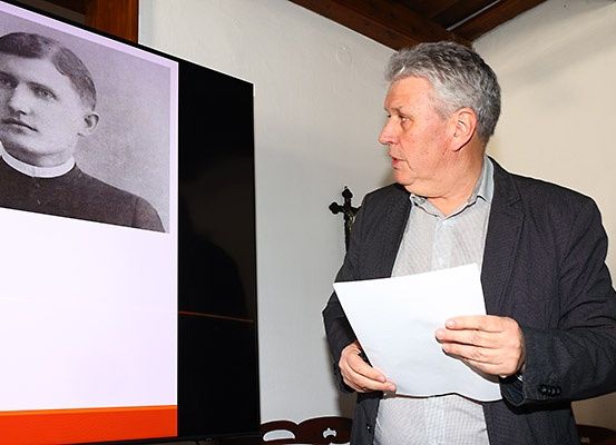 Ein Mann hält ein Papier in der Hand, während er vor einer Projektionsleinwand spricht, auf der ein altes Schwarz-Weiß-Foto eines jungen Mannes in geistlicher Kleidung zu sehen ist.
