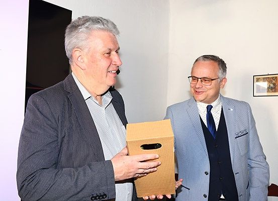 Zwei Männer in Anzügen tauschen lächelnde Blicke aus, während einer eine Schachtel hält, vor einer weißen Wand stehend.