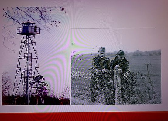 Ein Schwarz-Weiß-Bild von zwei Männern, die einen Stacheldrahtzaun reparieren, und einem Aussichtsturm.