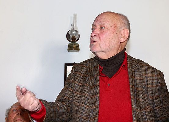 Ein älterer Mann in einem roten Hemd und einer karierten Jacke spricht, mit einer Öllampe und einem gerahmten Bild an der Wand hinter ihm.