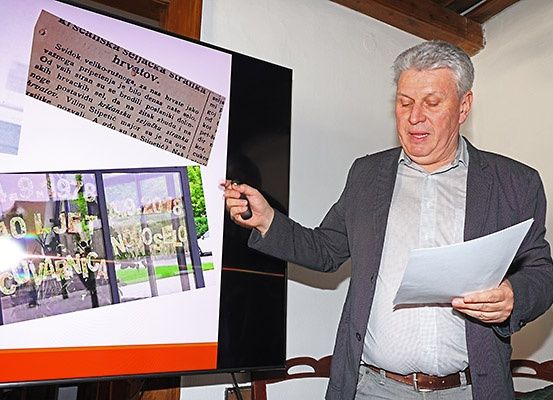 Ein älterer Mann hält eine Präsentation ab und zeigt auf einen Bildschirm, der ein Foto eines Gebäudes und einen Zeitungsausschnitt zeigt.