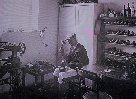 Ein Schuhmacher arbeitet in einer Werkstatt an einem Schuh mit einer Nähmaschine und Regalen voller Schuhe.