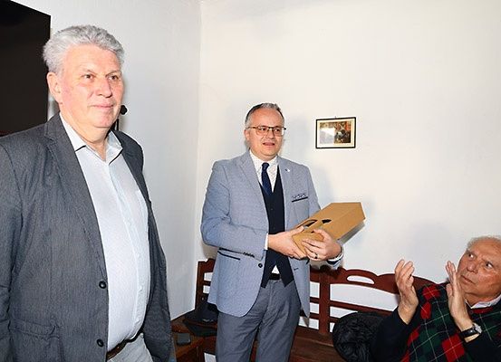 Drei Männer in Anzügen, einer hält eine Box, einer klatscht, stehen in einem Raum mit Holzstühlen und einem Bilderrahmen an der Wand.