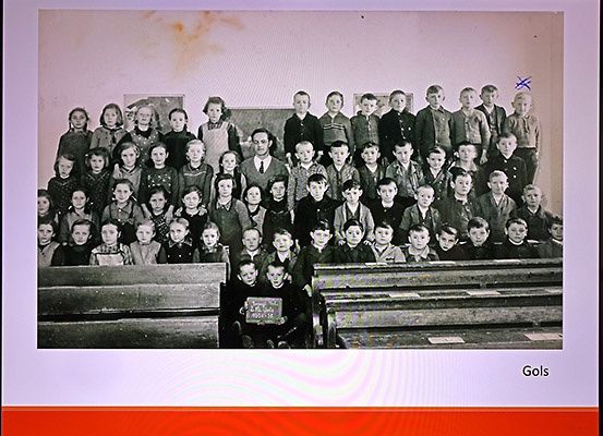 Ein Schwarz-Weiß-Foto eines Klassenzimmers mit Kindern und einem Lehrer, der davor steht. Sie tragen alle Uniformen und einige haben Namensschilder. Das Klassenzimmer hat Holzbänke.