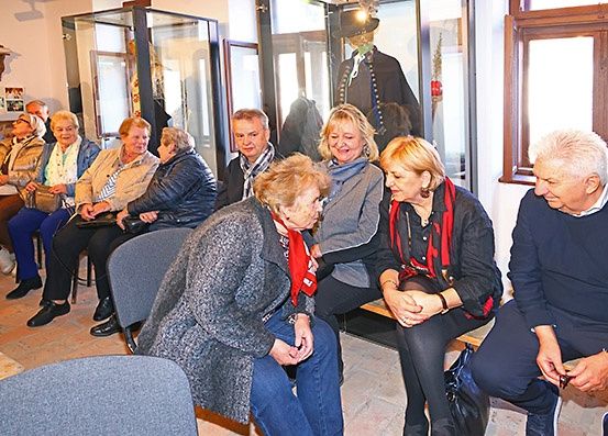 Mehrere ältere Menschen sitzen in einem Raum, wahrscheinlich in einem Museum, wobei sich eine Frau vorbeugt, um mit einer anderen zu sprechen. Sie scheinen in ein Gespräch vertieft zu sein.