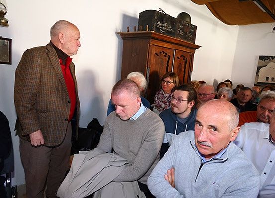 Mehrere Personen sitzen in einem Raum, einige mit Brille. Ein Mann steht mit einer Jacke über dem Arm. Ein hölzerner Schrank und eine Lampe sind an der Wand.