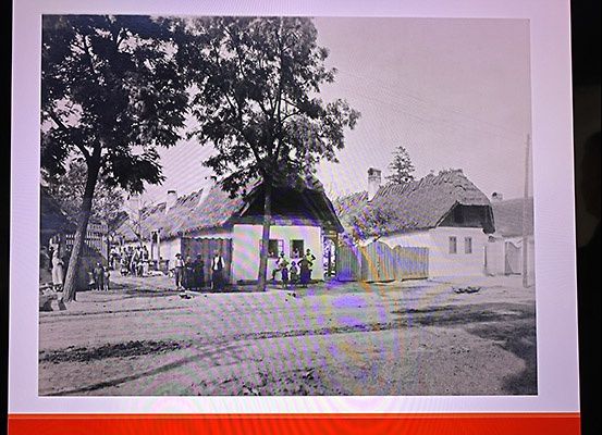Eine alte Schwarz-Weiß-Fotografie eines Dorfes mit einigen Menschen, die vor einem Haus mit Strohdach stehen.