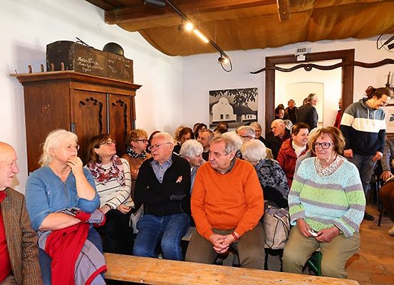 Eine Gruppe von Menschen sitzt auf Bänken in einem Raum mit Holzwänden, beleuchtet von Deckenleuchten. Eine Frau mit Brille trägt einen blauen Pullover.