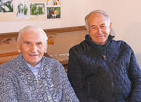 Zwei ältere Männer sitzen nebeneinander und lächeln. Sie tragen warme Kleidung. Hinter ihnen befindet sich eine Wand mit mehreren gerahmten Bildern.