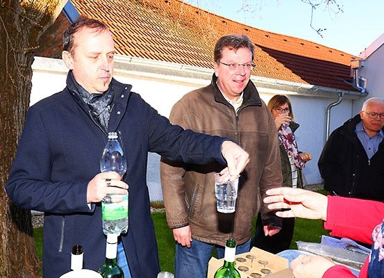 Mehrere Menschen stehen vor einem Haus. Ein Mann gießt Wasser aus einer Flasche in ein Glas. Ein anderer Mann lächelt.