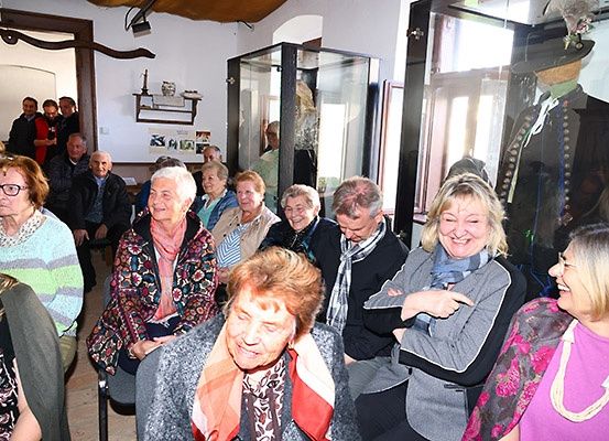 Eine Gruppe älterer Menschen sitzt in einem Raum, wahrscheinlich bei einem Treffen. Ein Dummy in traditioneller Kleidung ist in einer Glasvitrine ausgestellt.