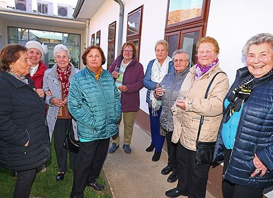 Eine Gruppe von Frauen in Jacken und Schals, einige mit Gläsern in der Hand, stehen in einer Reihe vor einem Gebäude. Sie lächeln und scheinen für ein Foto zu posieren. Das Gebäude hat Glasfenster und eine weiße Wand.