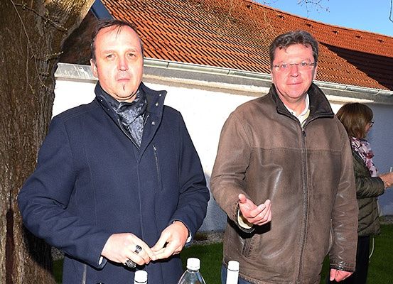 Zwei Männer stehen nebeneinander vor einem Haus mit einem roten Dach. Der Mann auf der linken Seite trägt eine blaue Jacke und einen Schal, während der Mann auf der rechten Seite eine braune Jacke und eine Brille trägt. Sie schauen beide nach vorne. Vor ihnen steht eine Flasche.