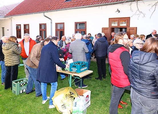 Eine Gruppe von Menschen, wahrscheinlich auf einer Veranstaltung, versammelt sich auf dem Rasen vor einem Gebäude. Einige stehen, andere sitzen und unterhalten sich. Ein Tisch mit Flaschen und einem Beutel ist vorhanden, und es ist ein klarer Himmel über ihnen.