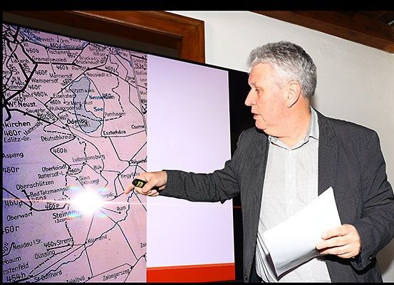 Ein älterer Mann präsentiert eine Karte auf einer Projektionsleinwand mit einem Laserpointer. Er trägt einen grauen Blazer und hält ein Papier in der anderen Hand.