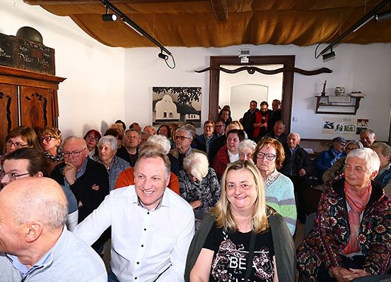 Eine Gruppe von Menschen sitzt in einem Raum, einige mit Brille. Ein Mann in einem weißen Hemd und eine Frau in einer blumigen Jacke lächeln. Es gibt einen Holzschrank und ein gerahmtes Bild an der Wand.