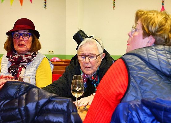 Drei Frauen in ausgefallenen Kostümen sitzen an einem Tisch und führen ein Gespräch. Sie befinden sich in einem Raum mit bunten Dekorationen.