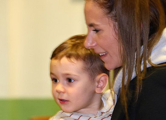 Eine Frau hält einen kleinen Jungen fest im Arm und schaut ihn lächelnd an. Der Junge sieht konzentriert aus, möglicherweise hört er zu oder schaut etwas an.