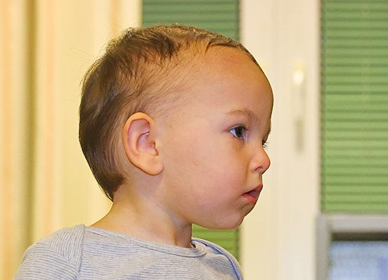 Ein kleiner Junge mit kurzen Haaren und blauen Augen schaut nach oben. Er trägt ein graues Shirt und steht drinnen. Hinter ihm befindet sich ein Fenster mit grünen Jalousien und einem weißen Rahmen.