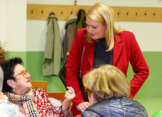 Eine Frau in einem roten Blazer spricht mit einer älteren Frau, die eine Brille und einen Schal trägt, in einem Raum mit grünen und weißen Wänden.