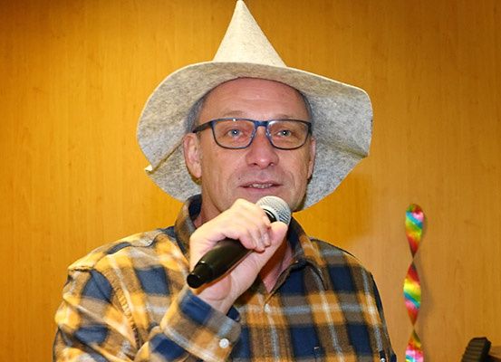 Ein Mann mit einem breitkrempigen Hut und kariertem Hemd spricht in ein Mikrofon. Er trägt eine Brille und steht vor einer Holzwand.