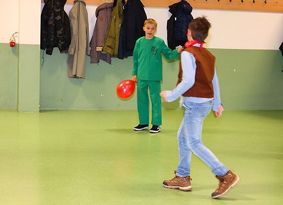 Zwei junge Jungen spielen mit einem roten Ballon in einem Raum mit grünem Boden. Ein Junge trägt ein grünes Outfit, während der andere eine braune Weste trägt.