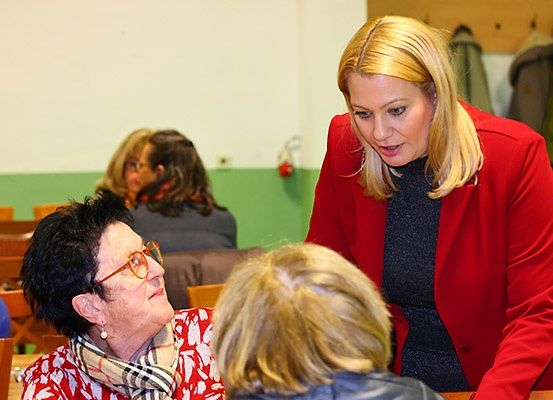 Eine Frau in einem roten Blazer unterhält sich mit einer älteren Frau, die eine Brille und ein Halstuch trägt, während eine weitere Frau zusieht. Der Raum ist grün und weiß gestrichen, mit Luftballons und Jacken im Hintergrund.