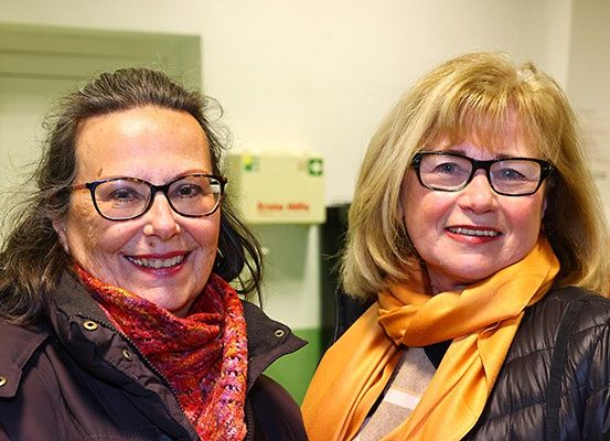 Zwei Frauen mit Brille und Schals, eine mit einem roten Schal und die andere mit einem gelben Schal, lächeln für ein Foto in einem Innenbereich.