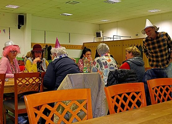 Eine Gruppe von Menschen mit Partyhüten und lässiger Kleidung, die an einem Tisch sitzen, wobei ein Mann in der Nähe eines Holzschranks steht.