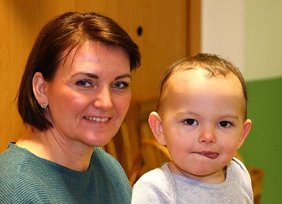 Eine Frau und ein Kind lächeln in die Kamera und stehen eng beieinander in einem Raum mit Holz- und grünen Wänden.