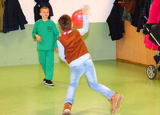 Zwei Jungen spielen mit einem roten Ballon in einem Raum mit grünen Wänden und an der Wand hängenden Kleidern.