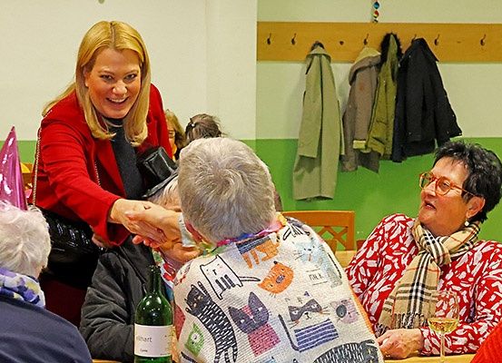 Mehrere ältere Frauen sitzen an einem Tisch, eine von ihnen trägt ein katzenthematisches Lätzchen. Eine Frau in einem roten Mantel gibt einer anderen die Hand. Mäntel hängen an der Wand.