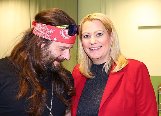 Ein Mann mit langen Haaren, der ein rotes Stirnband und eine Sonnenbrille trägt, steht neben einer Frau in einem roten Mantel. Sie lächeln beide für die Kamera.
