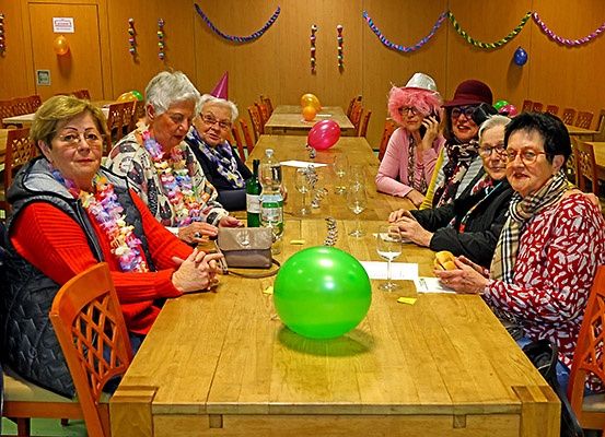 Eine Gruppe älterer Frauen feiert an einem festlich gedeckten Tisch mit Luftballons und Dekorationen, trägt Hüte und Schals, lächelt und posiert für ein Foto.