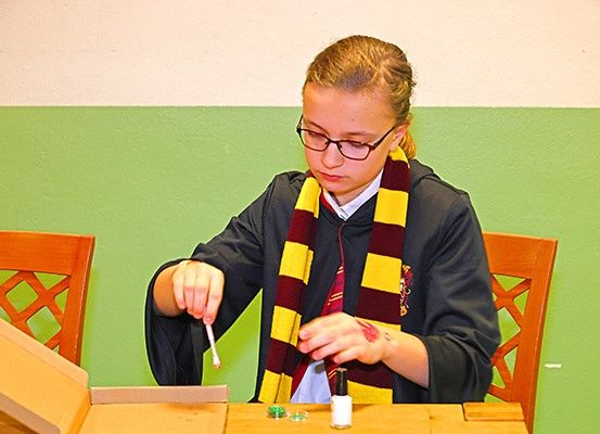 Ein junges Mädchen in einem Gryffindor-Umhang sitzt an einem Tisch, hält eine Zauberstäbe und Farbe in der Hand, mit einer grün-weiß gestreiften Wand im Hintergrund.