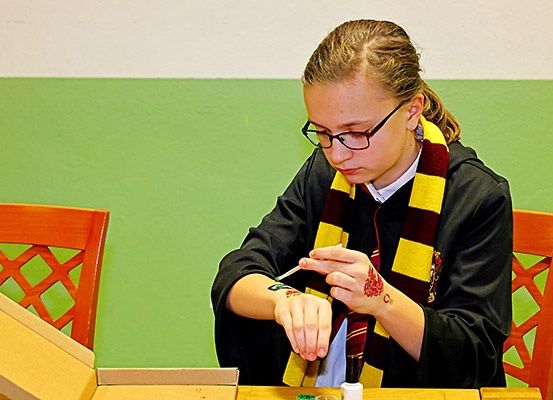 Eine junge Frau sitzt an einem Tisch, trägt einen Gryffindor-Umhang und eine Schürze. Sie malt ein rotes Design auf ihren Arm.