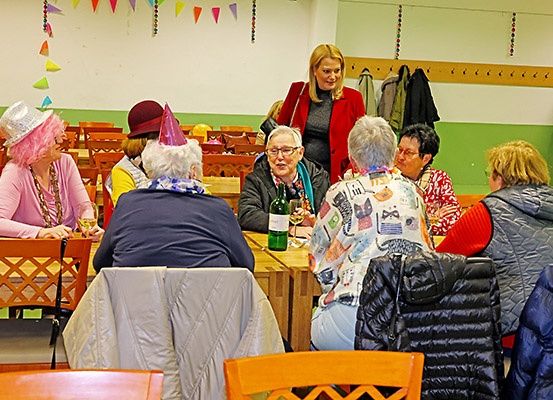Eine Gruppe älterer Frauen in einem Raum, einige sitzen um einen Tisch herum, während eine steht und lächelt. Sie tragen Hüte und einige haben Brillen. Der Raum ist mit Flaggen und einem Regal mit Jacken dekoriert.