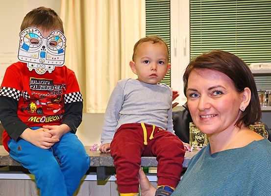 Zwei Kinder und eine Frau sitzen auf einem Tresen in einem Raum. Ein Kind trägt ein rotes Shirt mit Autoaufdruck und das andere ein graues Shirt und rote Hosen. Die Frau lächelt.